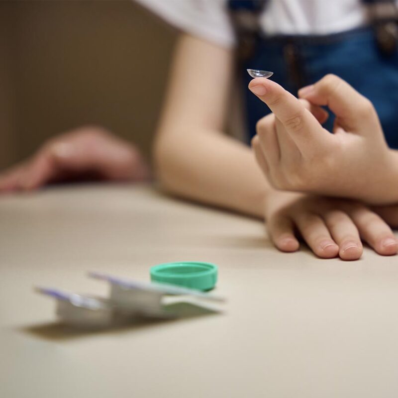 Girl holds a transparent contact lens on her finger, a child in denim overalls